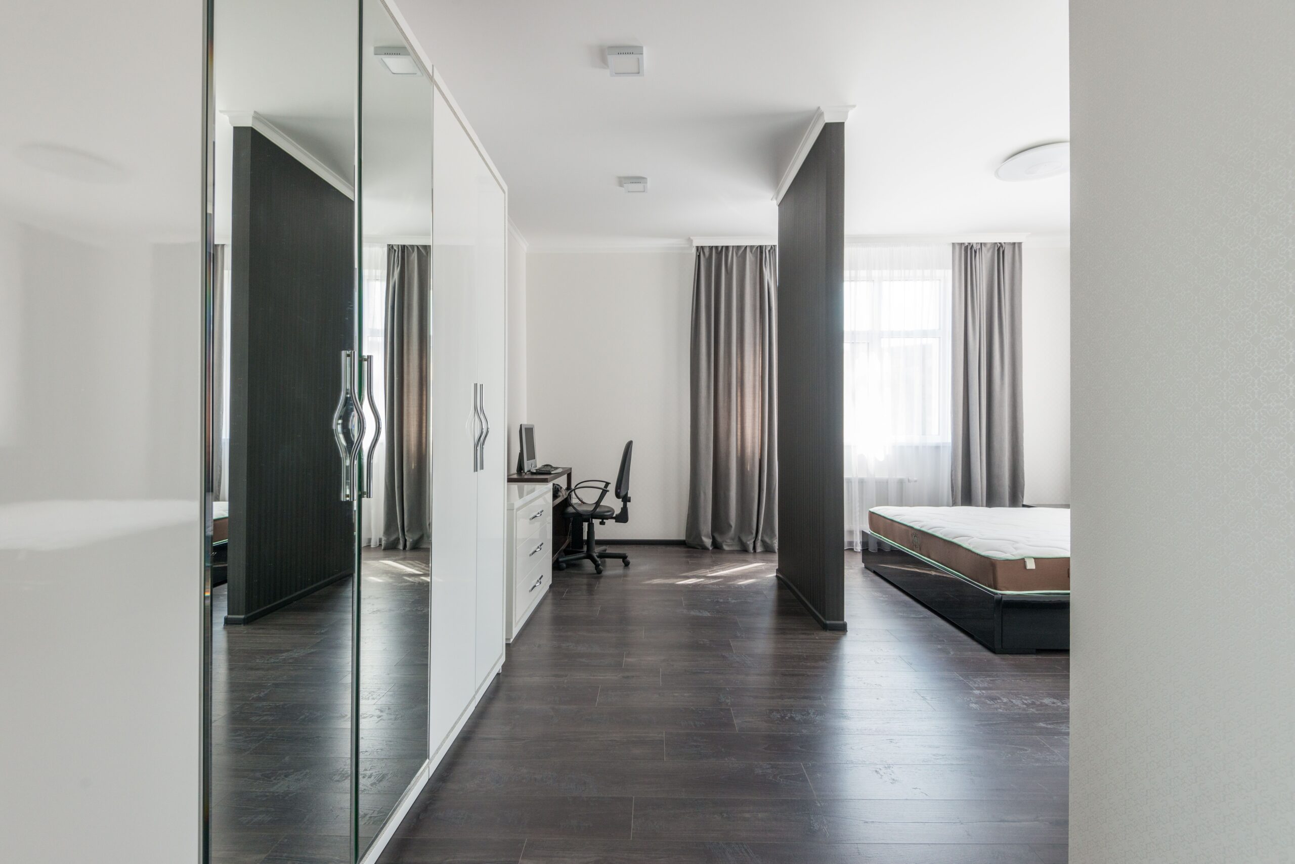 Modern minimalist bedroom with dark wood floors, large mirrored closet, desk and chair, bed without bedding, and windows with gray curtains letting in natural light.